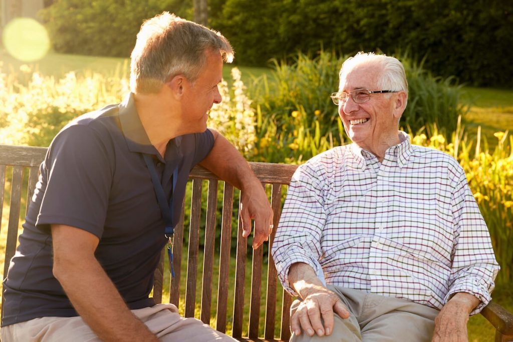 Adult son sitting outside on a bench with senior father smiling & talking together