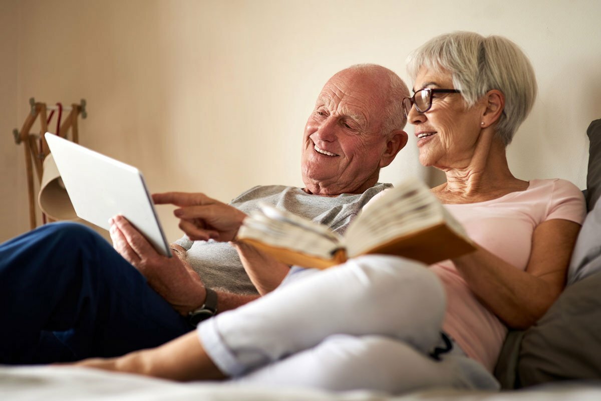 Older couple laying in bed reading and browsing a tablet