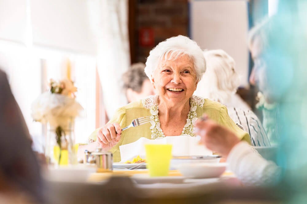 Smiling senior woman with gray hair sitting in The Buckingham's dining hall enjoying a meal with friends.