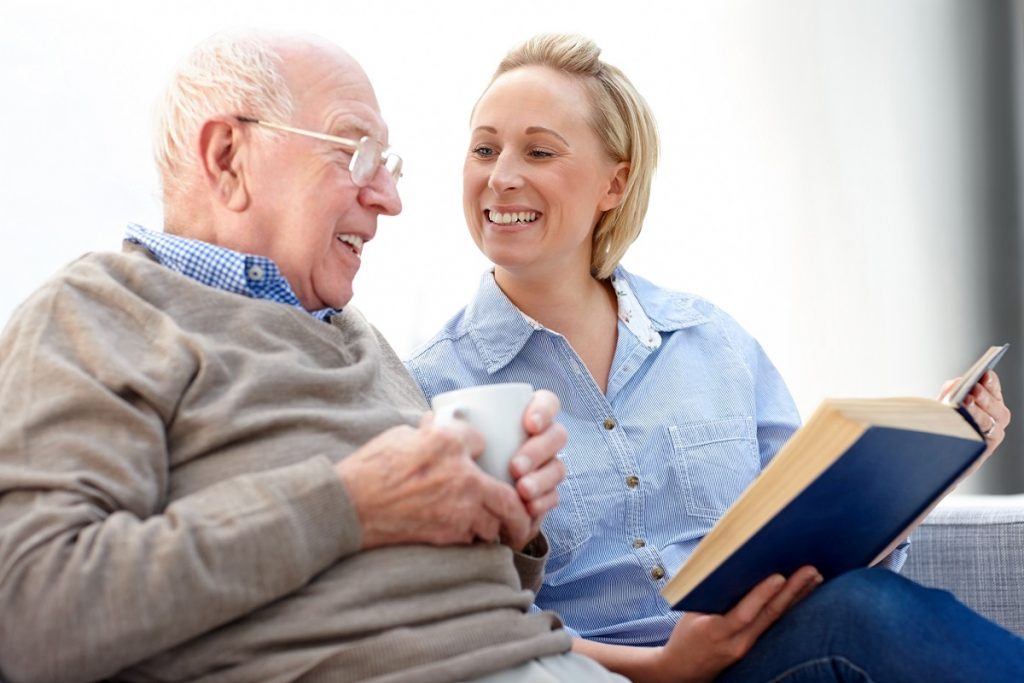 A happy senior man with female caregiver reading a novel while sitting together on sofa.